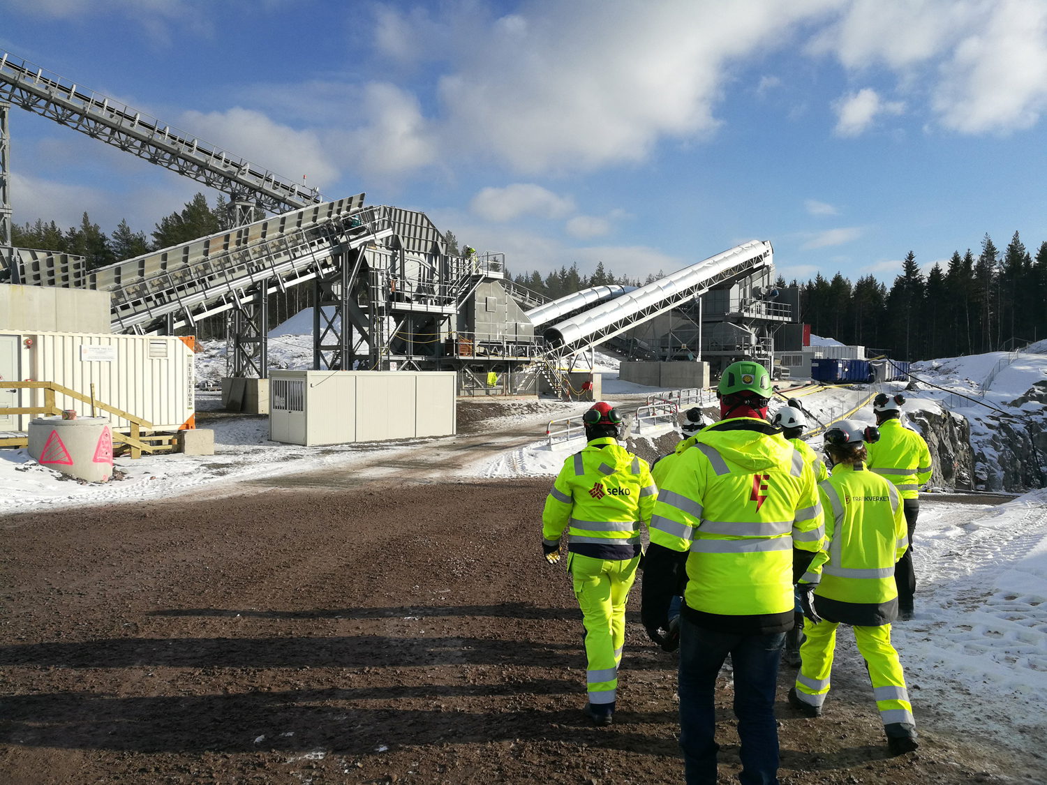 BIlden föreställer sex personer i gula reflexjackor som är på väg mot stenkrossen, en stor industrikonstruktion med bandgångar och baracker. Personerna har bygghjälmar på sig, varav en har en grön skyddsombudshjälm och ELektrikerförbundets symbol på sin jacka. En annan person har symbolen för Seko på sin jacka.