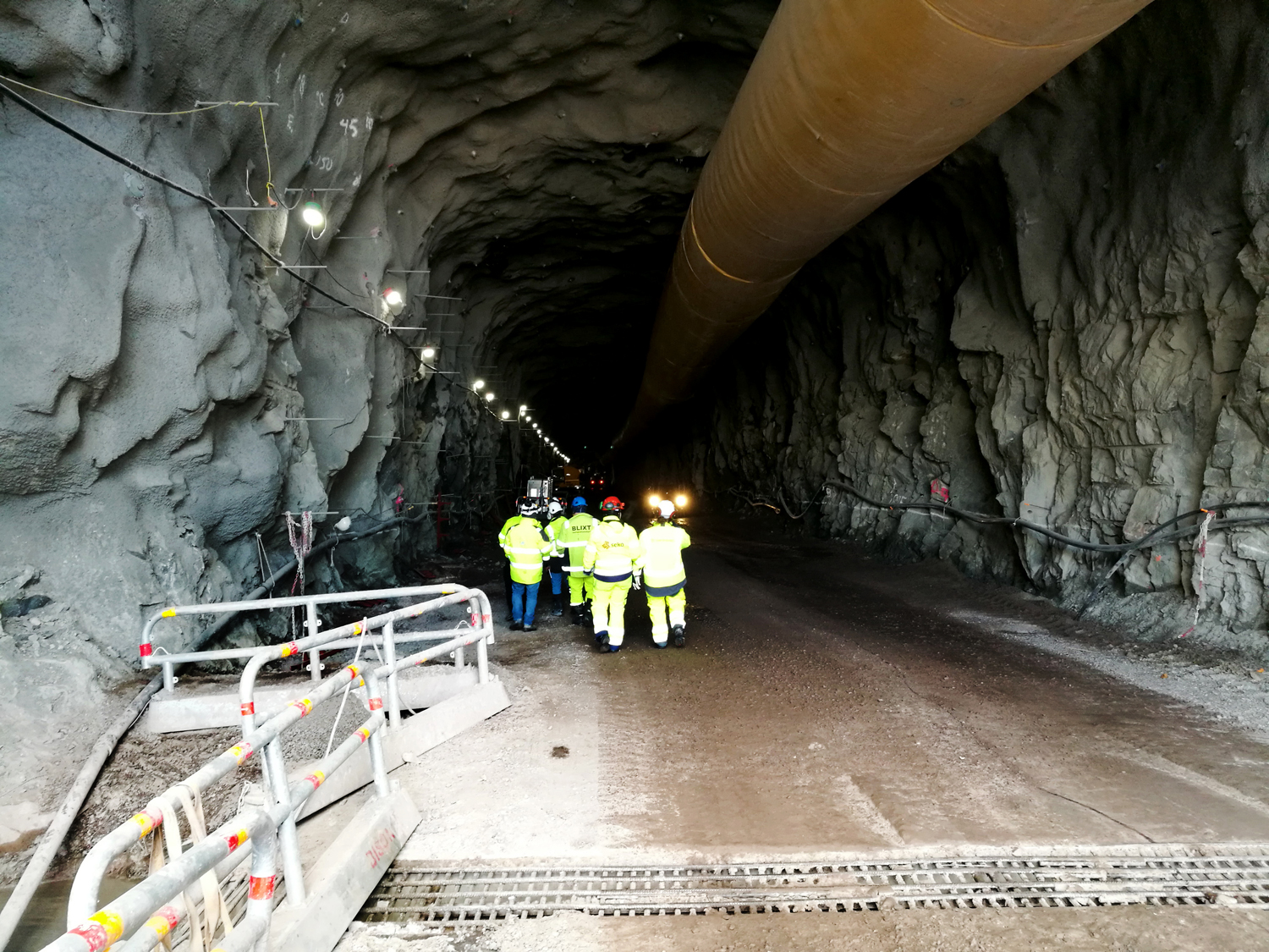 BIlden föreställer ryggarna på en grupp personer i gula reflexjackor, som är på väg in i en mörk tunnel. I tennelns tak löper tii stort rör och i förgrunden på bilden syns avspärrningsräcken i metall.