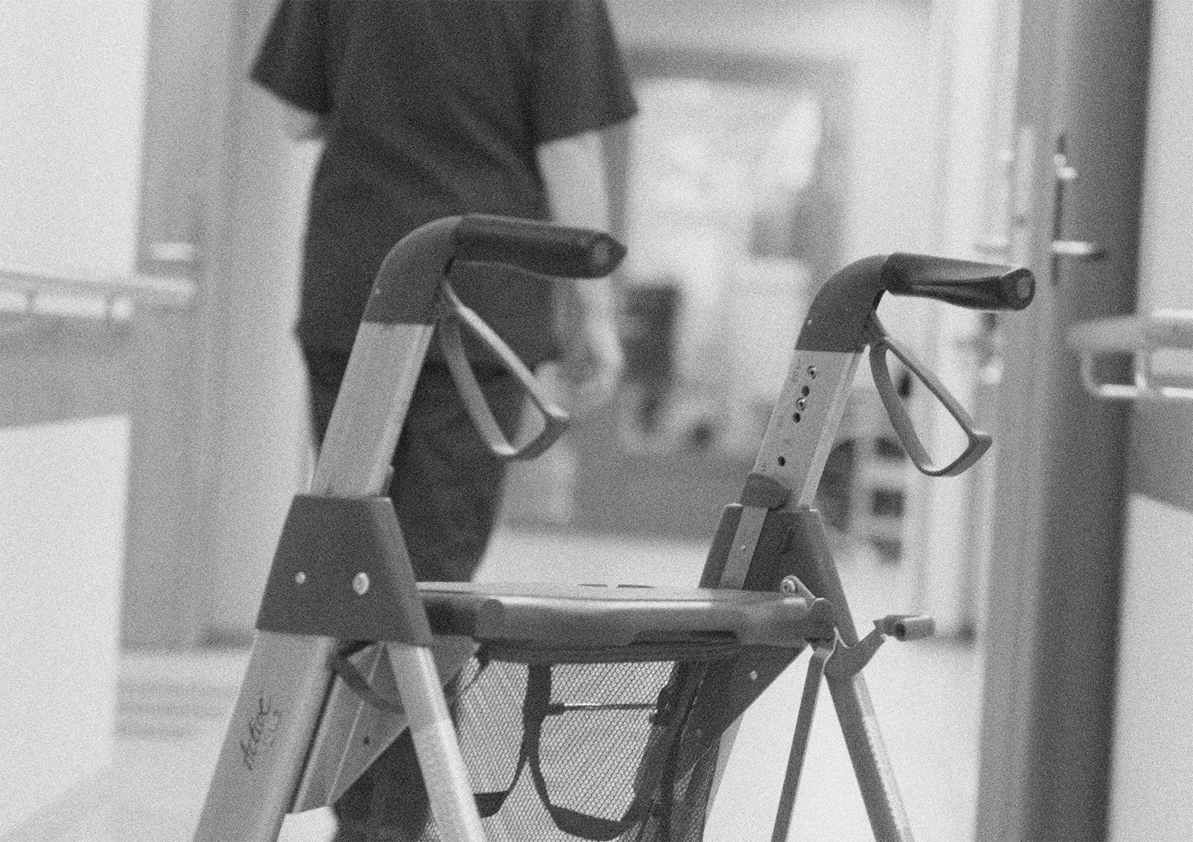 En undersköterska går i en korridor. I förgrunden syns en rollator.