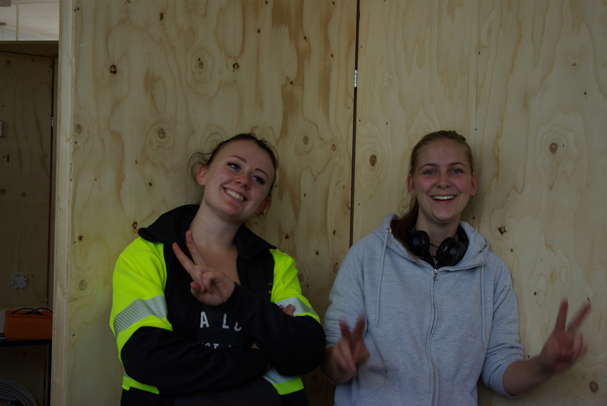 Två glada personer står framför en plywood-vägg. En med armarna i kors och en gör peace-tecknet.
