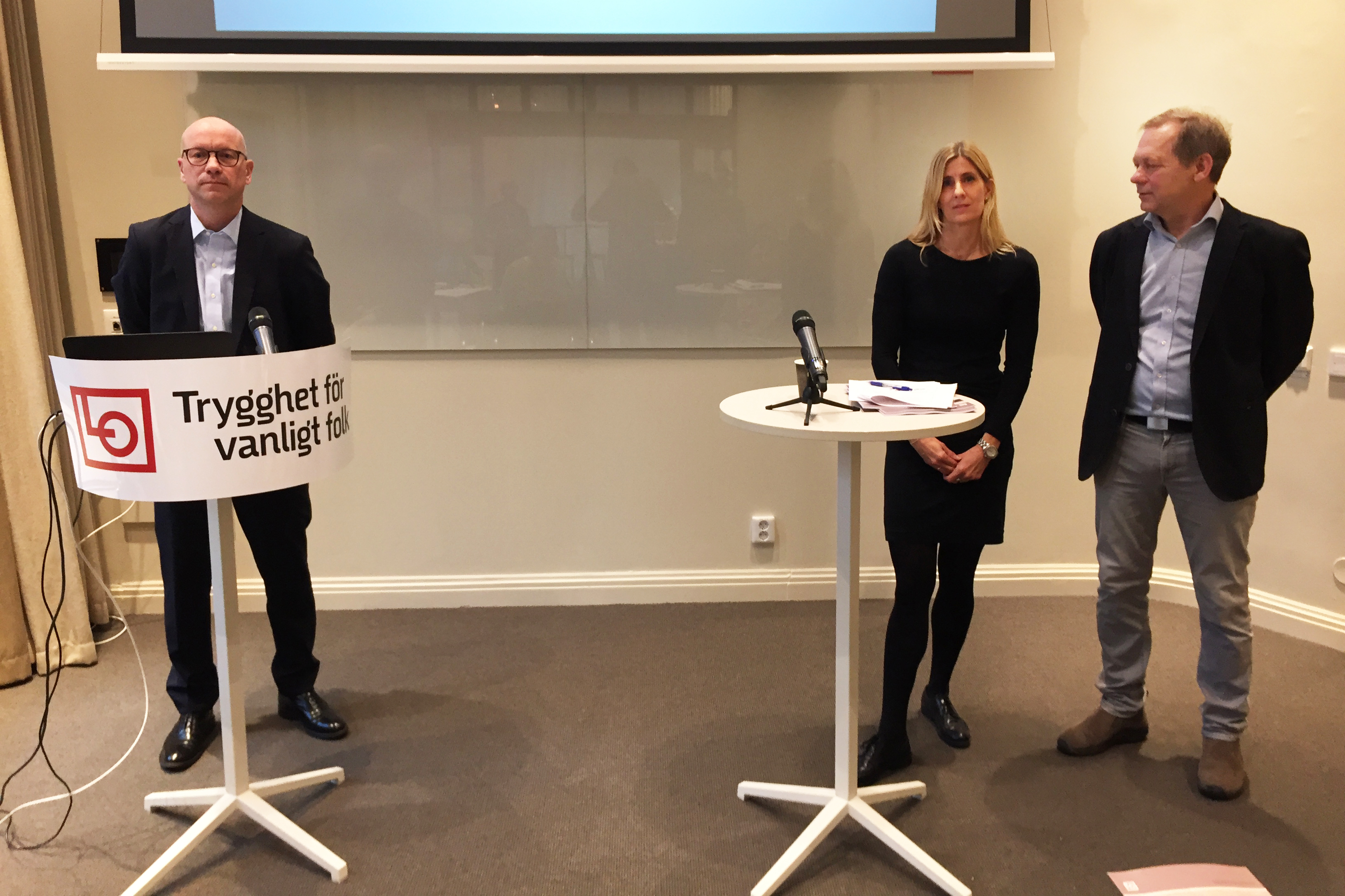 Tre personer står vid två talarbord framför en tom whiteboard. Ett av borden har en banderoll med LO:s logotyp på.