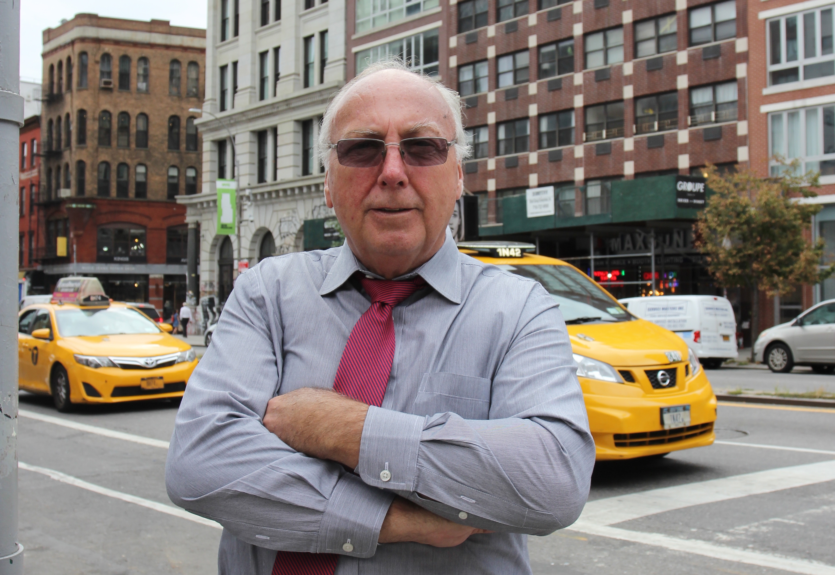 Chris Erikson står på ett övergångsställe med en stadsvy i bakgrunden, bland annat två gula taxibilar.