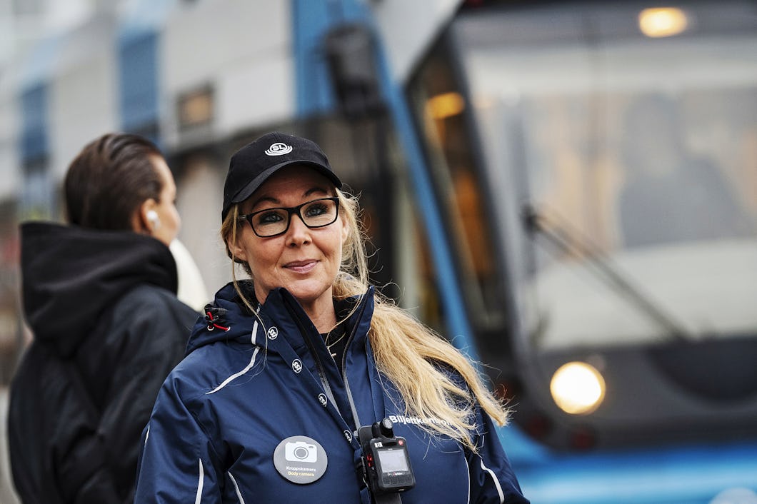 Ivana i uniform står vid en spårvagnshållplats och håller en enhet i handen. En spårvagn syns i bakgrunden.