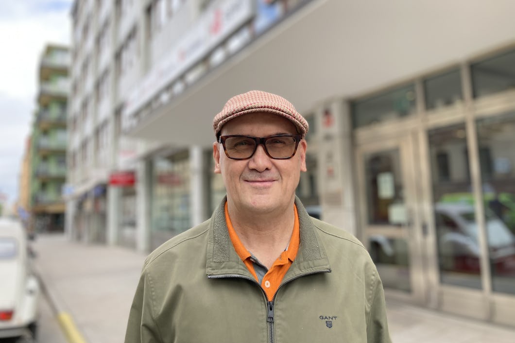 En man med glasögon, keps och jacka står på en trottoar framför en byggnad med glasdörrar.