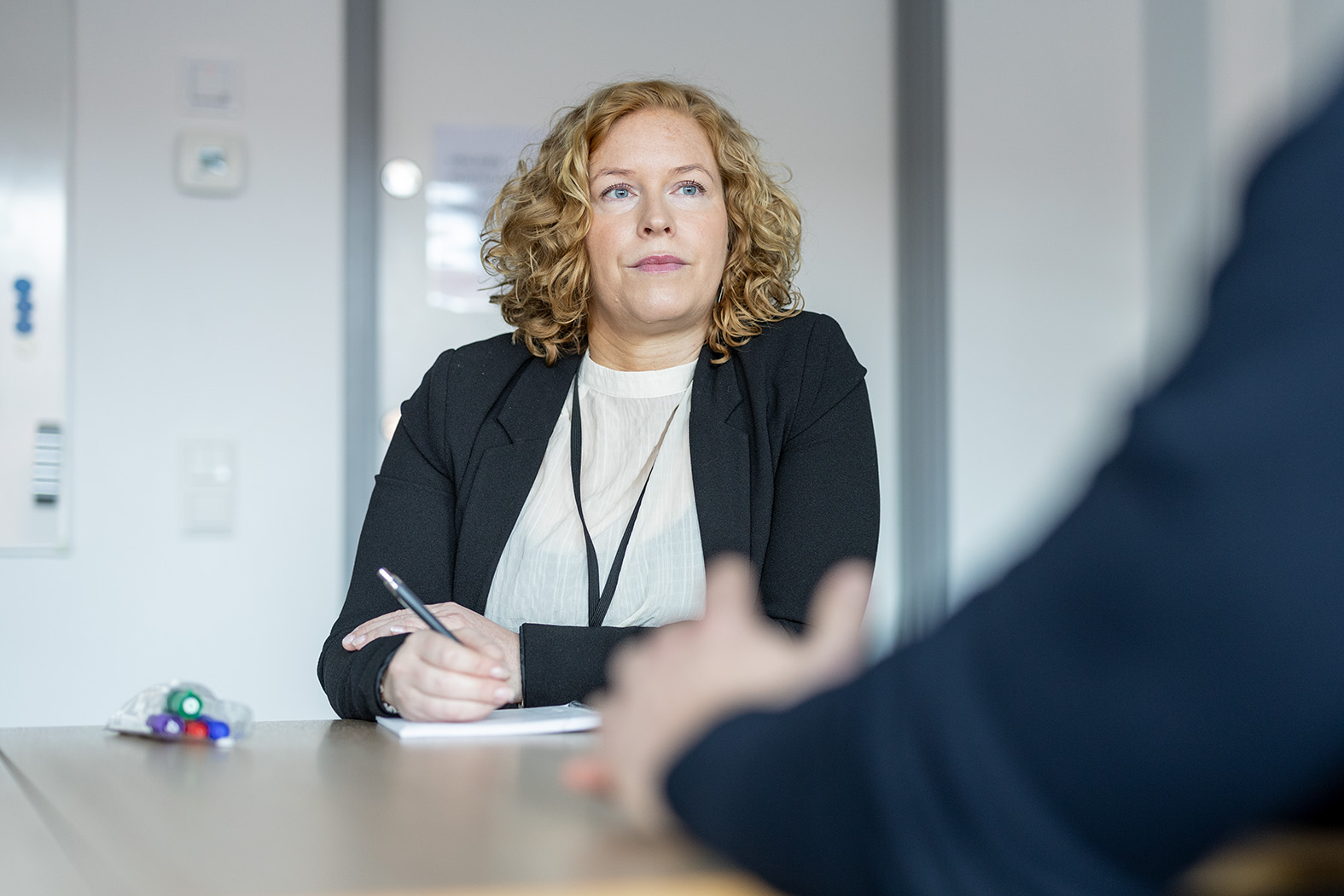 Bild på blond kvinna i vit tröja oc h mörk kavaj. Hon sitter vid ett bord och antecknar något. Bakom henne vita väggar, en vit dörr, och kanten på en whiteboard. I förgrunden någon annans suddiga vänsterarm.