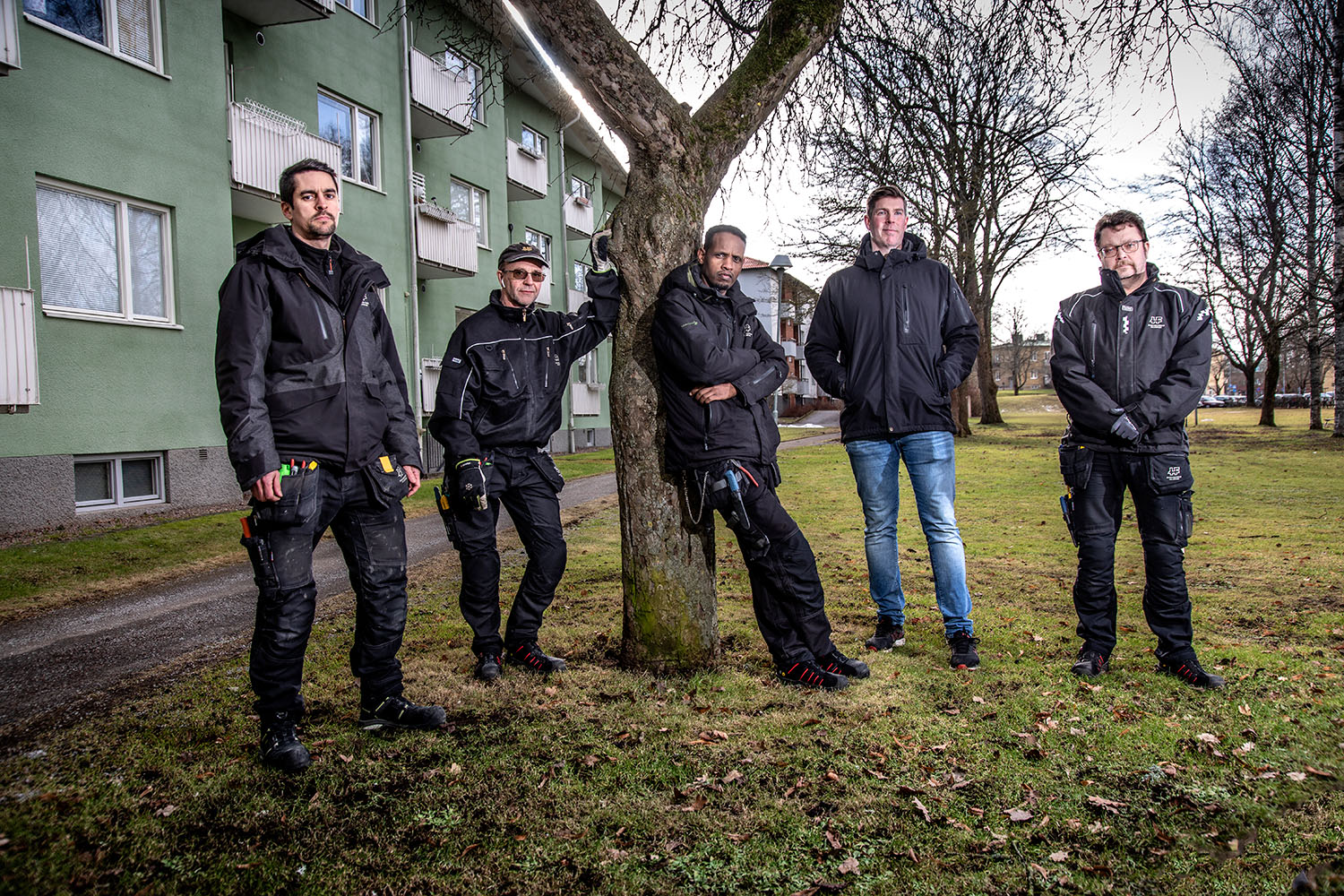 Fem män i arbetskläder står vid ett träd i ett bostadsområde. Alla tittar mot kameran.
