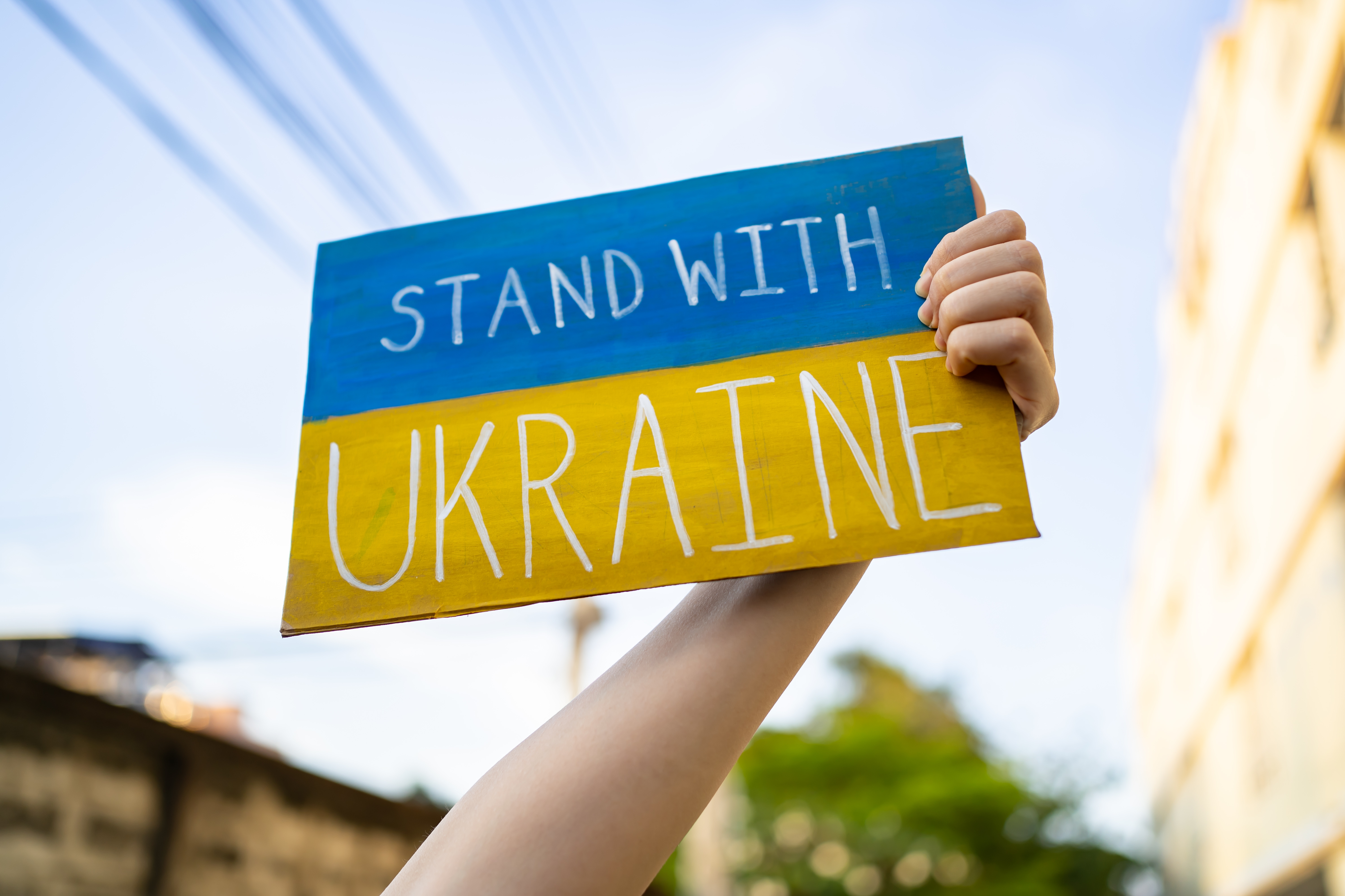 En hand håller upp en skylt med Ukrainas flagga, samt texten: "Stand with Ukraine."