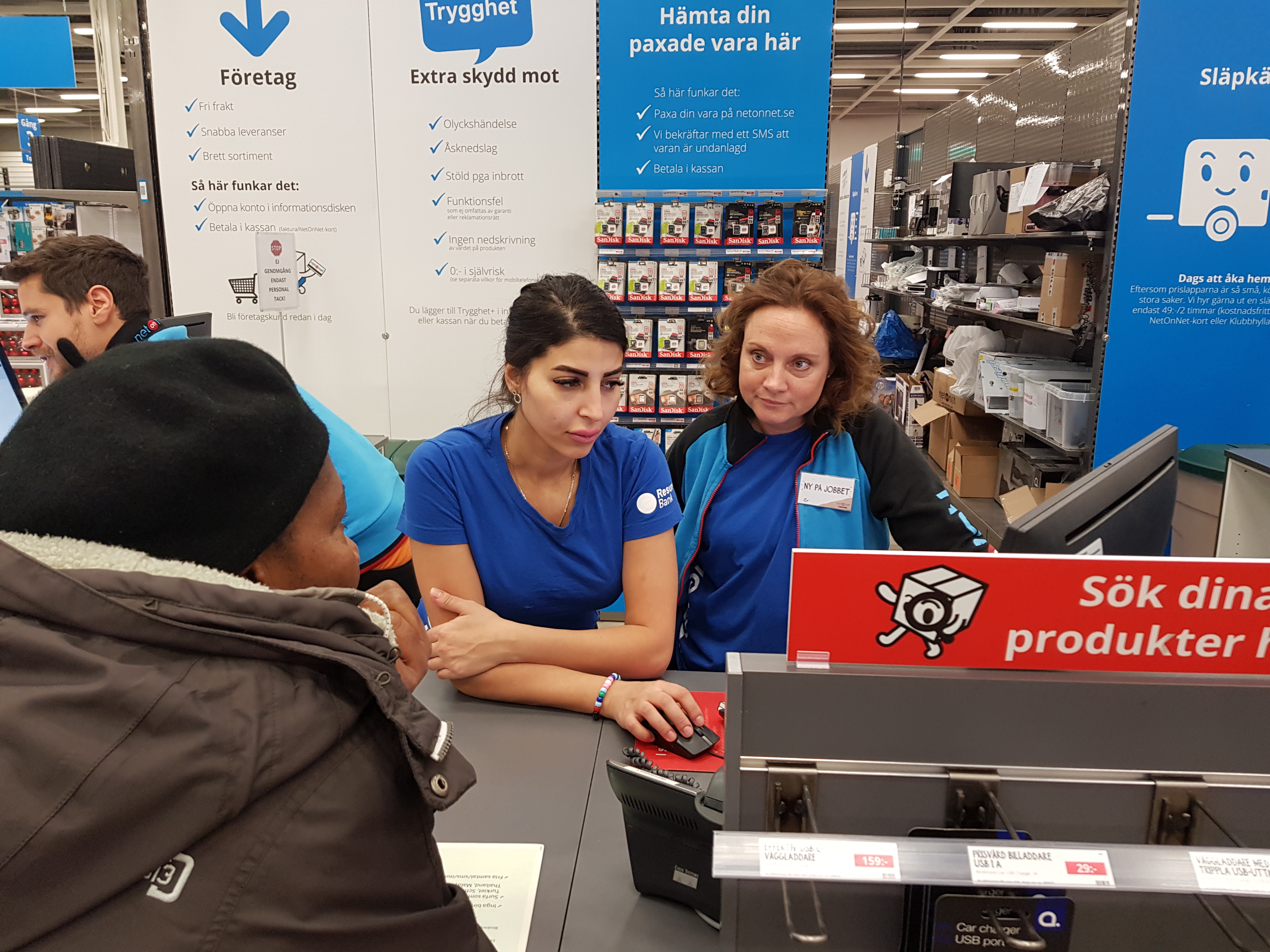 Två kvinnor bakom disken hjälper en kund. Bakom dem syns lagorhyllor och skyltar som upplyser kunderna.