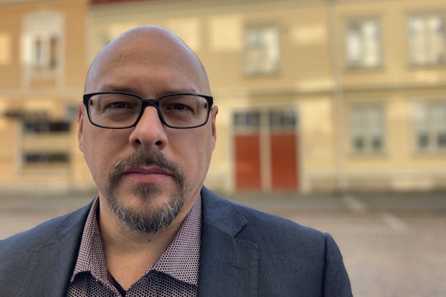 Anders Fennsjö står utomhus, iklädd en kavaj och mönstrad skjorta. I bakgrunden syns en gul byggnad med vita fönsterkarmar och röda dubbeldörrar.