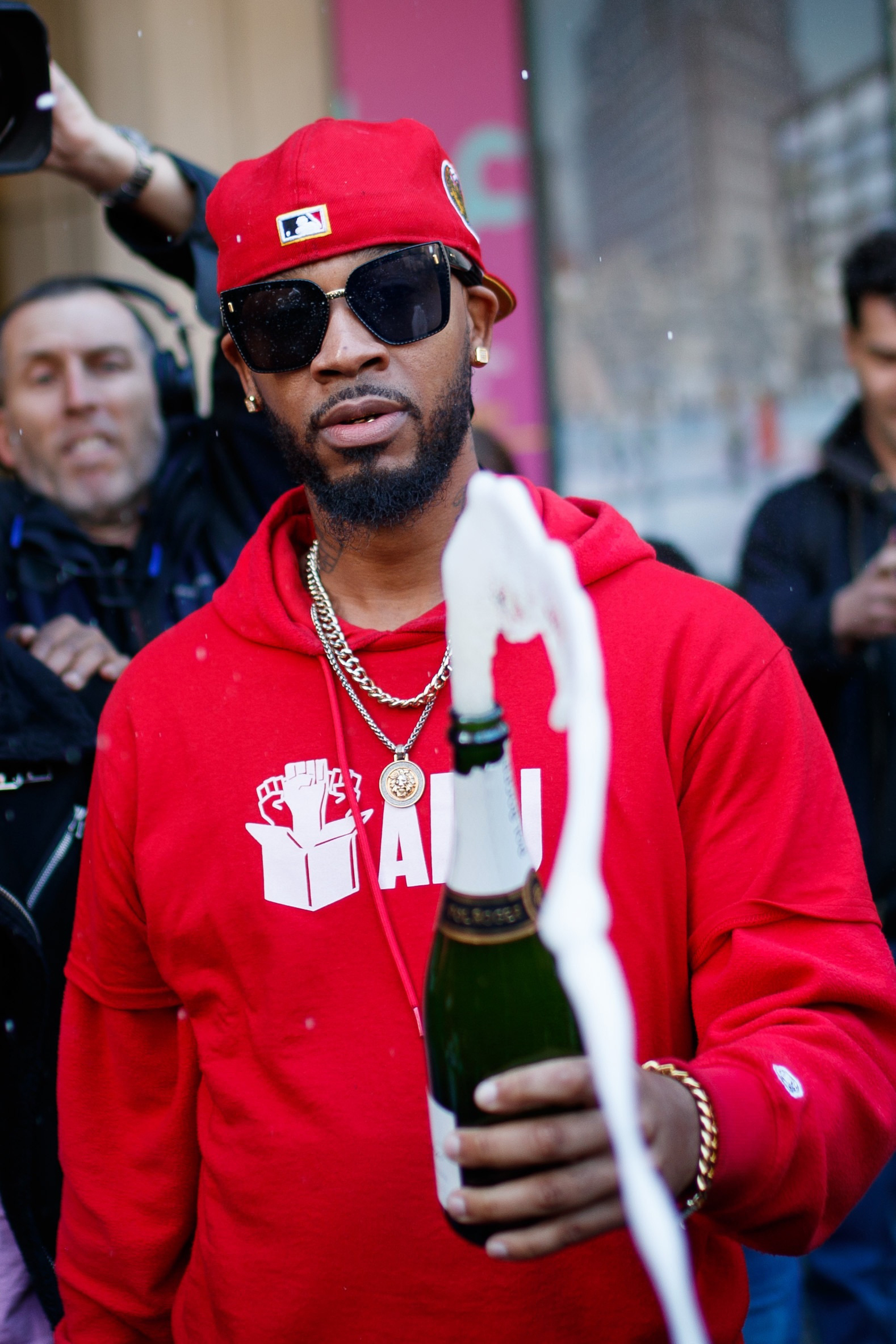 Chris Smalls, iklädd bakåtvänd röd keps, röd tröja och stora svarta solglasögon, lyfter en champagneflaska i en skål, bubblor sprutar ur flaskan.