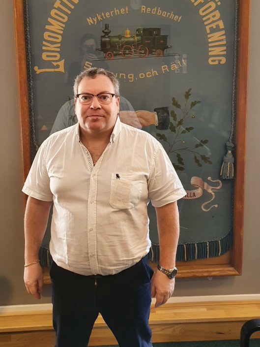 En man i vit skjorta och mörka byxor står framför en tavla med ett gammal lok på.