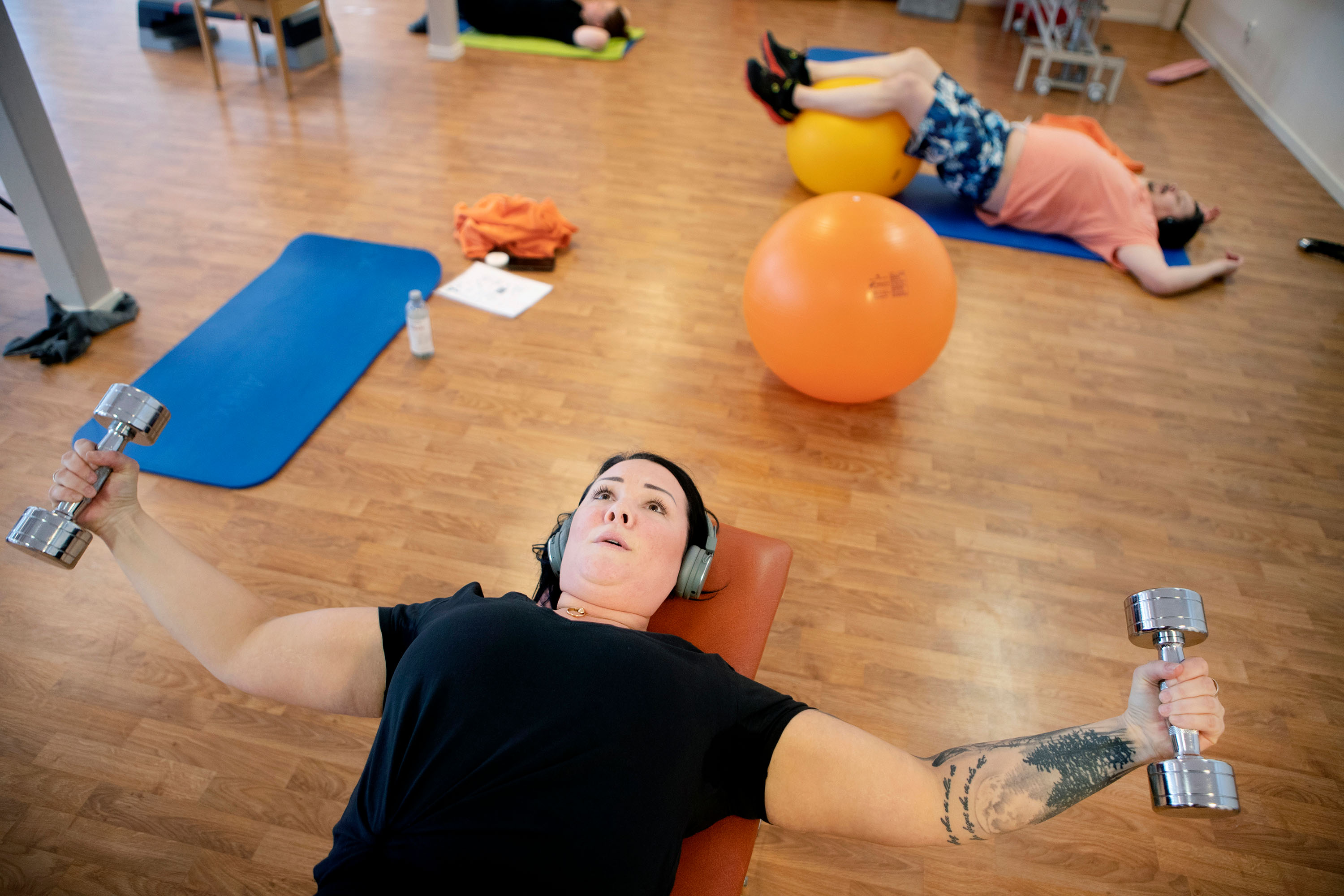 Längst fram i bilden en kvinna i halvfigur, hon lyfter ett par hantlar. I bakgrunden tränar två personer till, och runt dem ligger olika träningsredskap.