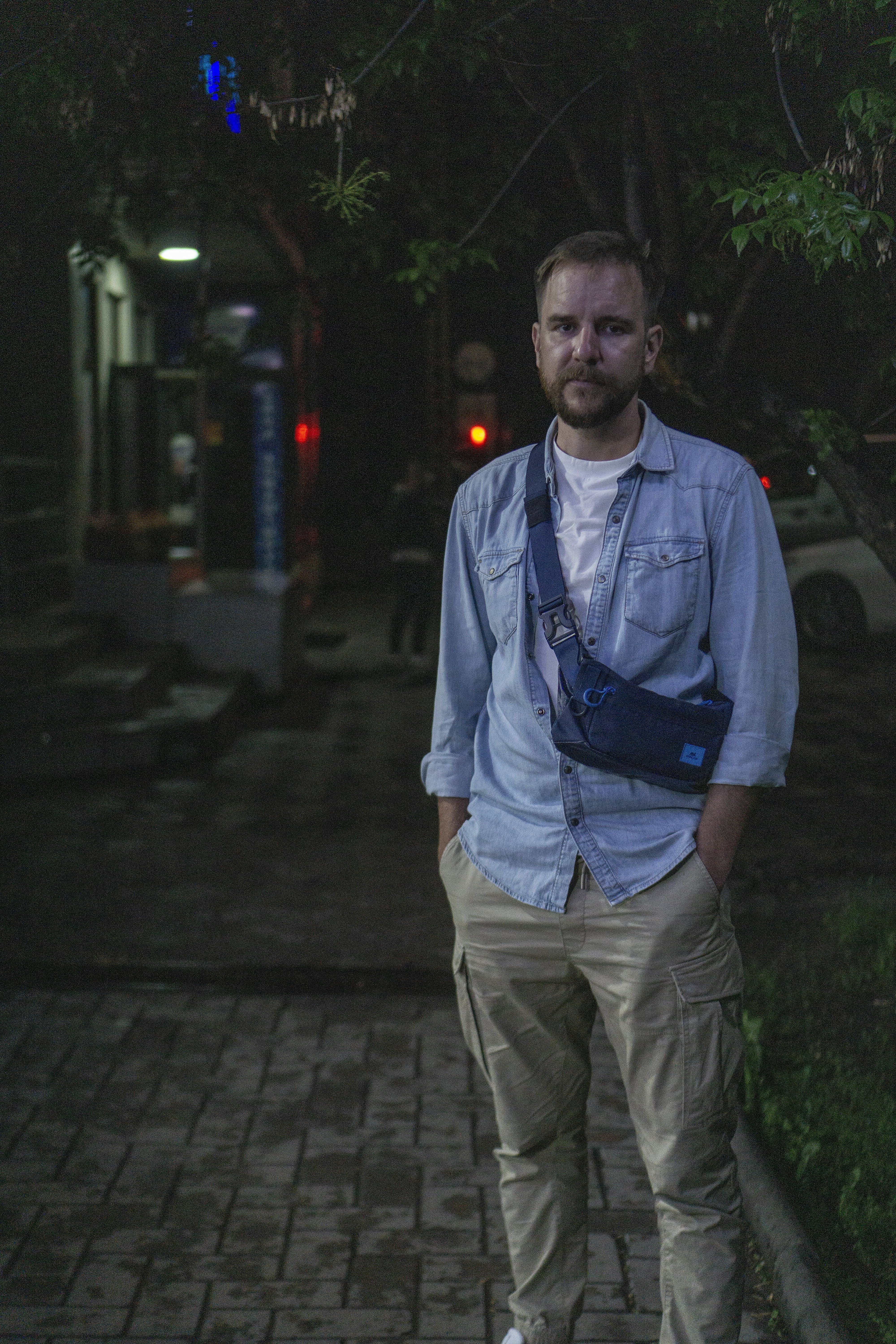En man i jeansskjorta med uppkavlade armar och ljusa khakibyxor står på en mörk gata. Han har kortklipp brunt hår, kort skägg och en väska hängande över magen.