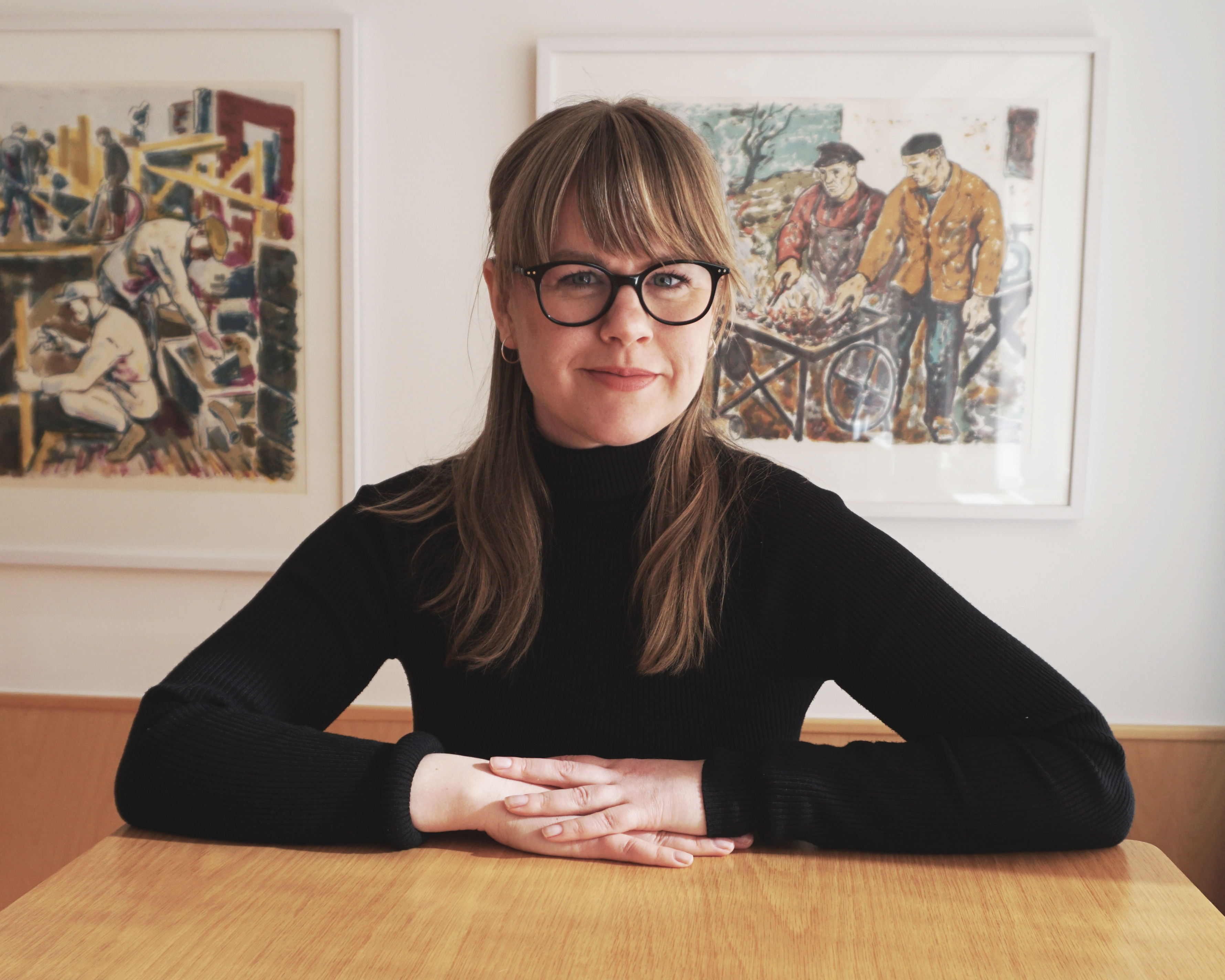 Eleonore Eriksson sitter vid ett bord, bakom henne hänger två tavlor.