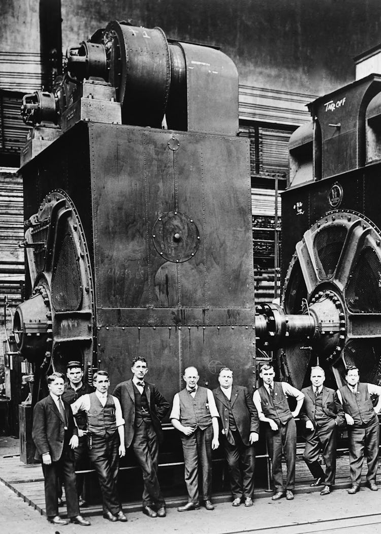 Nio kostymklädda män poserar framför två massiva elektriska generatorer i en fabrik. Svartvitt foto, cirka 1920.