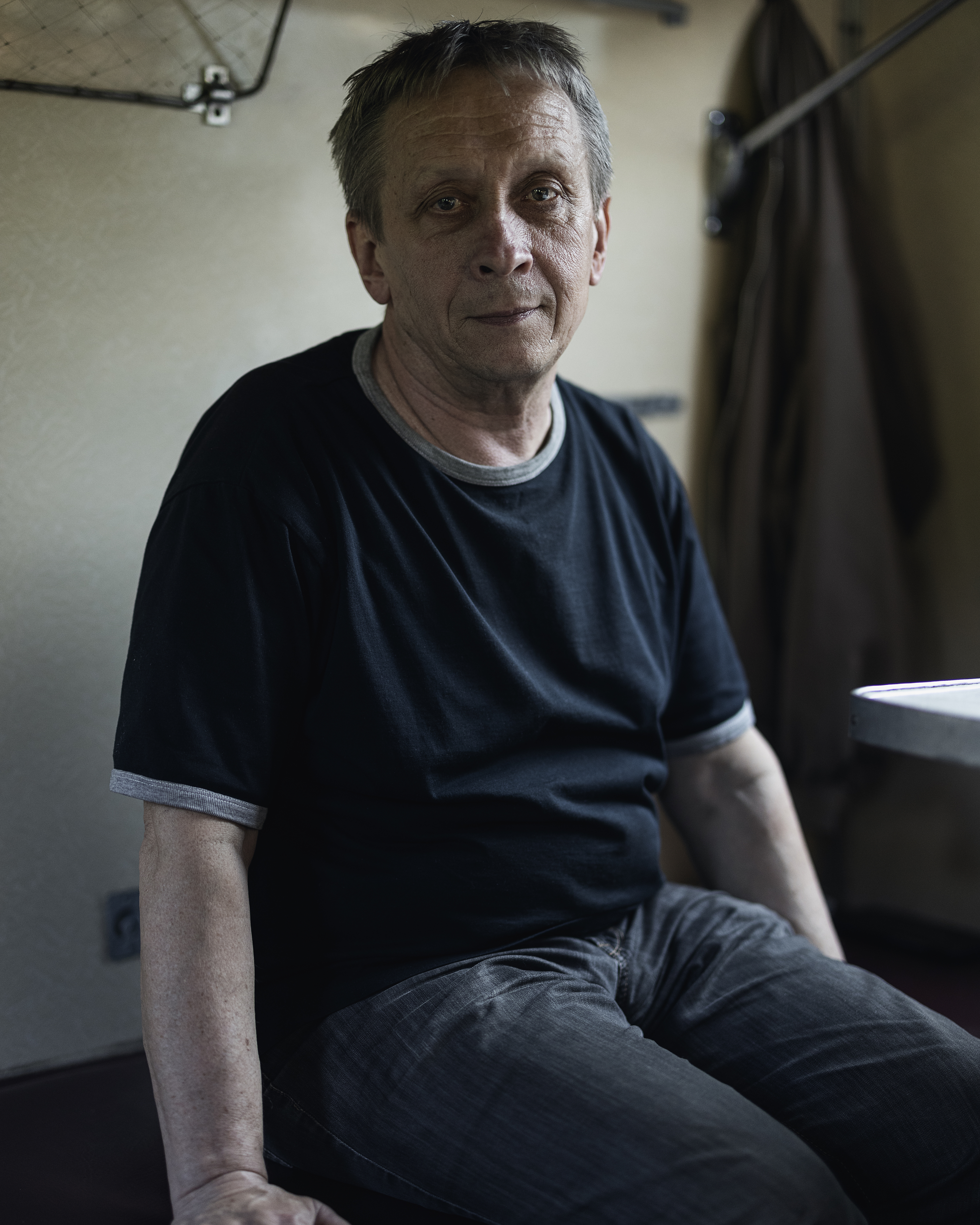 En gråhårig medelålders man i svart t-tröja och mörka jeans sitter på ett tågsäte och tittar mot kameran.