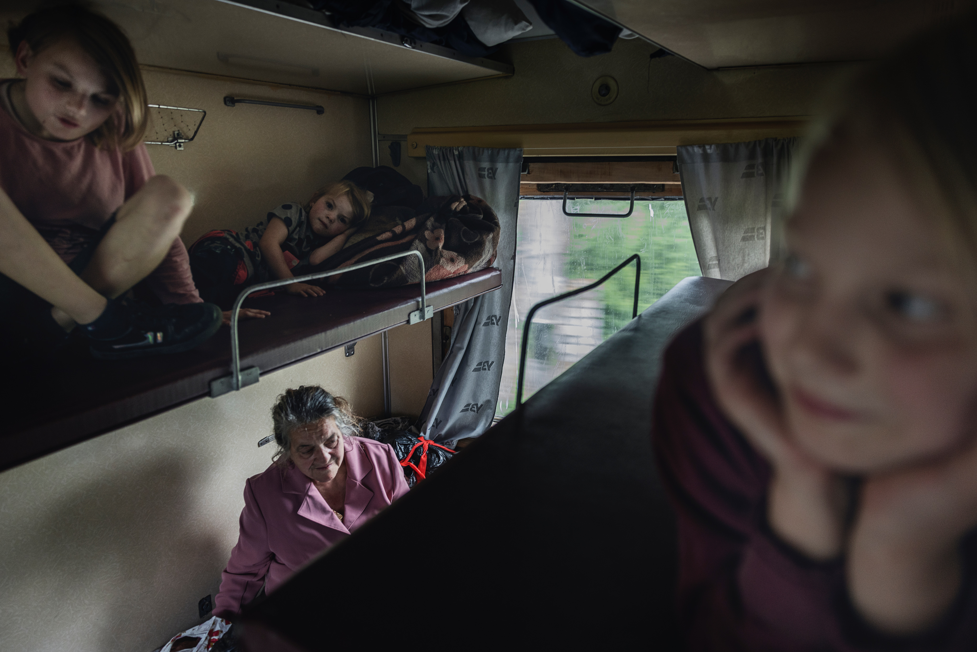 Tre blonda barn sitter på varsin överslaf i en tågkupé. På britsen nedtill syns en äldre gråhårig kvinna i Rosa kappa.