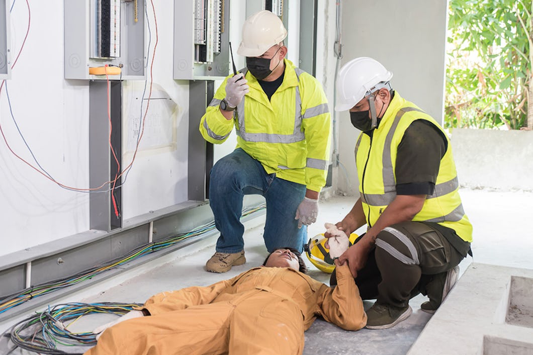 Två män i skyddsväst och hjälm hjälper en tredje man som ligger på golvet, till synes medvetslös. De finner sig precis vid elektrisk utrustning.