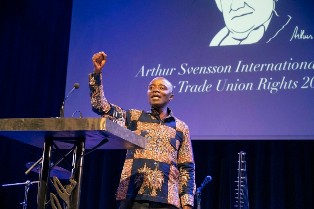 George Poe Williams står på en scen framför en blå bakgrund. Han bär traditionella liberiska kläder och höjer ena näven i en kampgest.