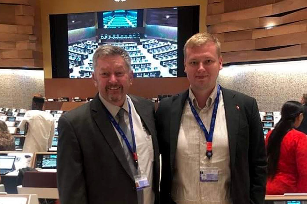 Två män i kostym, en äldre mörkhårig och en yngre blond, står axel mot axel i ett konferensrum och tittar mot kameran.