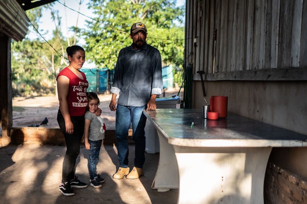 A man, a woman and child stands in the shade of a tin roof.