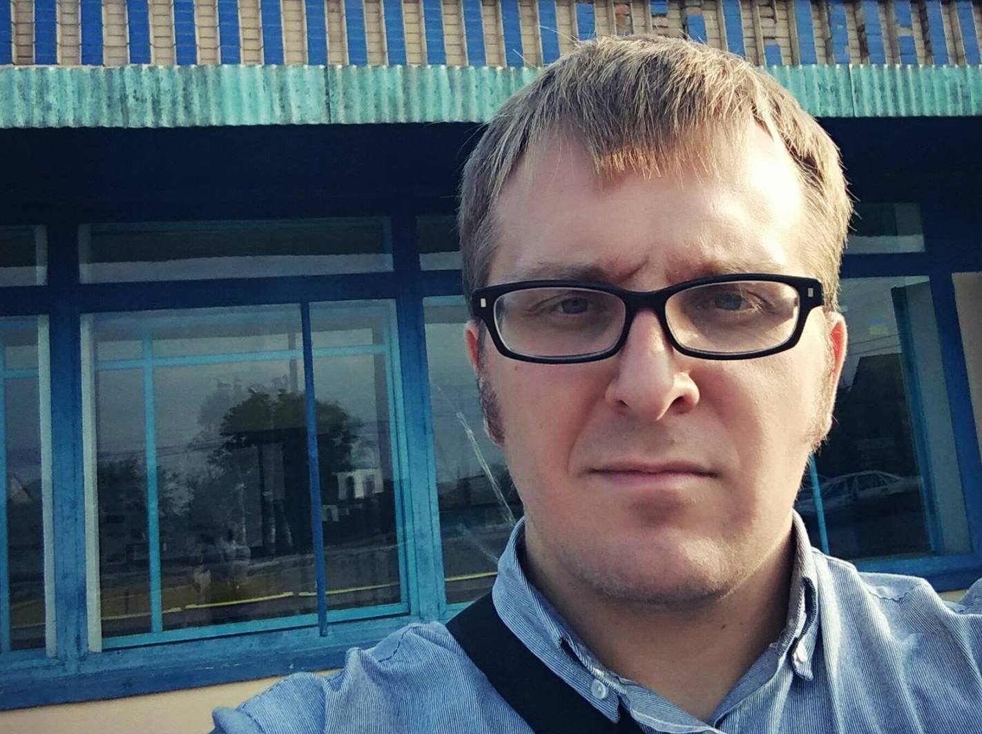 Närbild på en blond man med svarta glasögon och neutralt ansiktsuttryck.