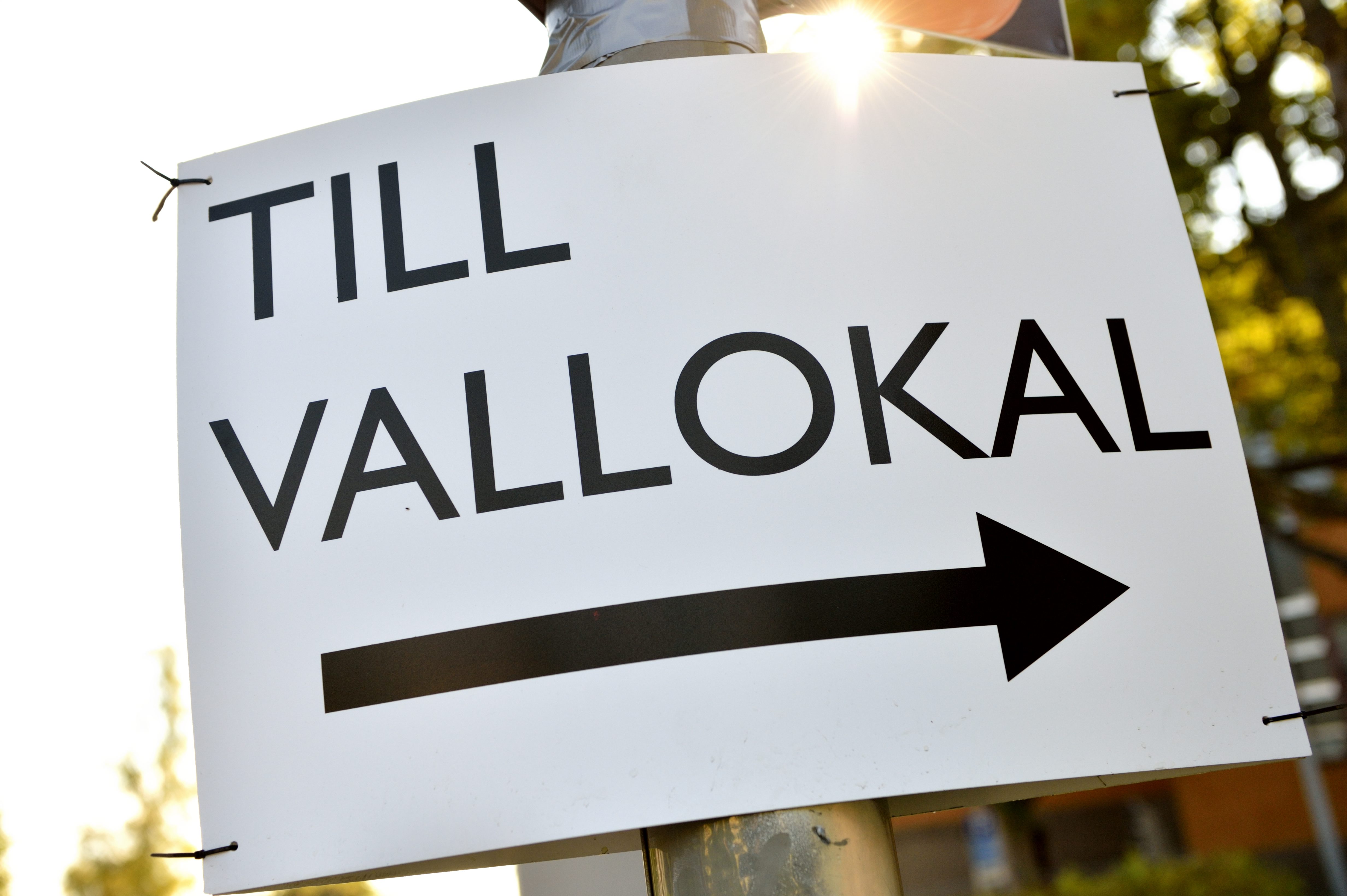 En vit skylt med en svart pil och texten "Till vallokal" hänger på en stolpe.