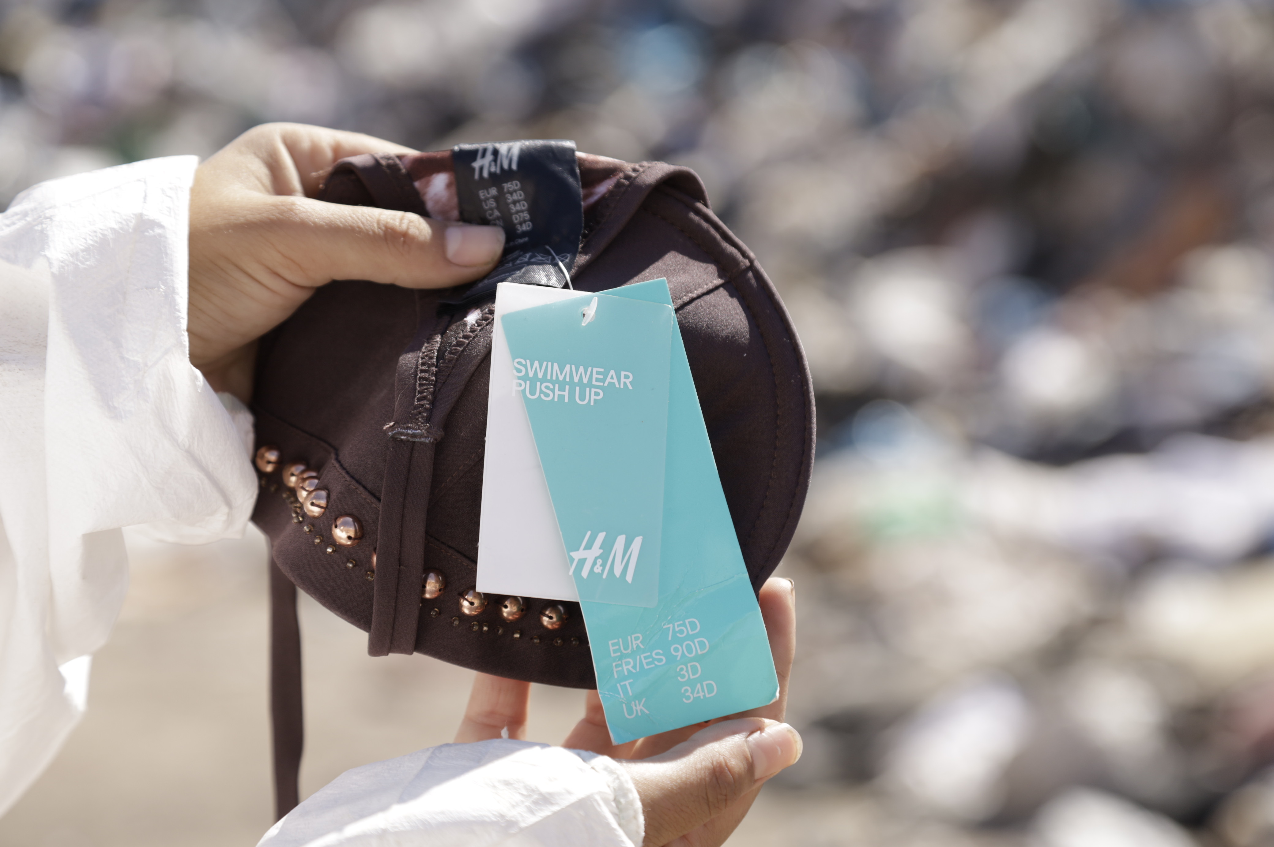 En person som inte syns i bild, förutom armarna, håller upp ett plagg från H&M framför en hög med dumpade kläder.