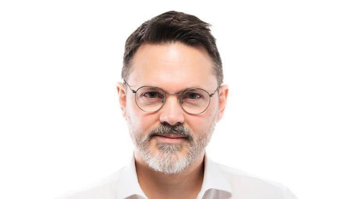 En man med kort mörkt hår och skägg, som bär runda glasögon och en vit skjorta, mot en enkel vit bakgrund.