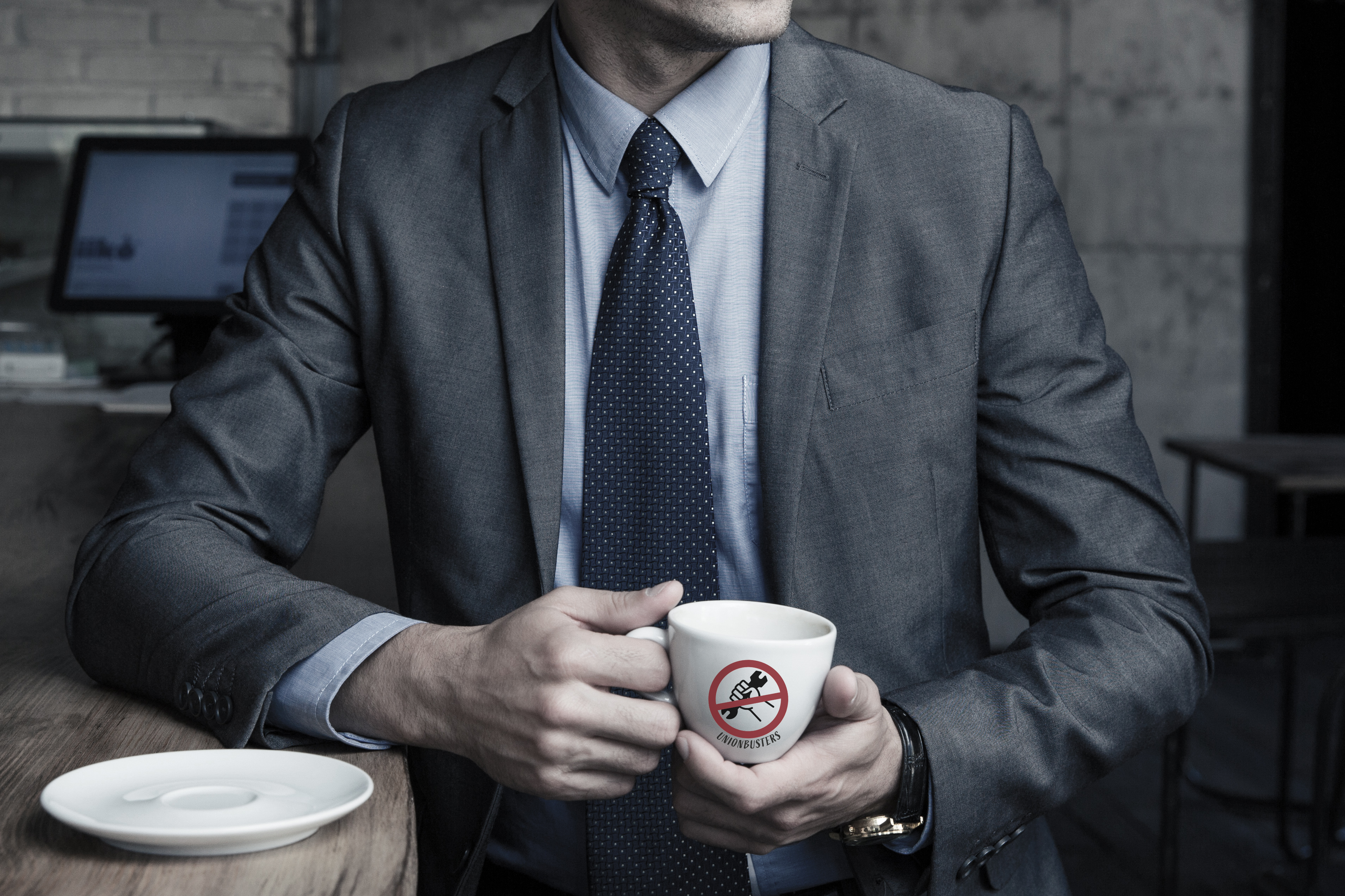 En man i grå kostym, vars ansikte inte syns, håller en kaffekopp med texten "Union busters".