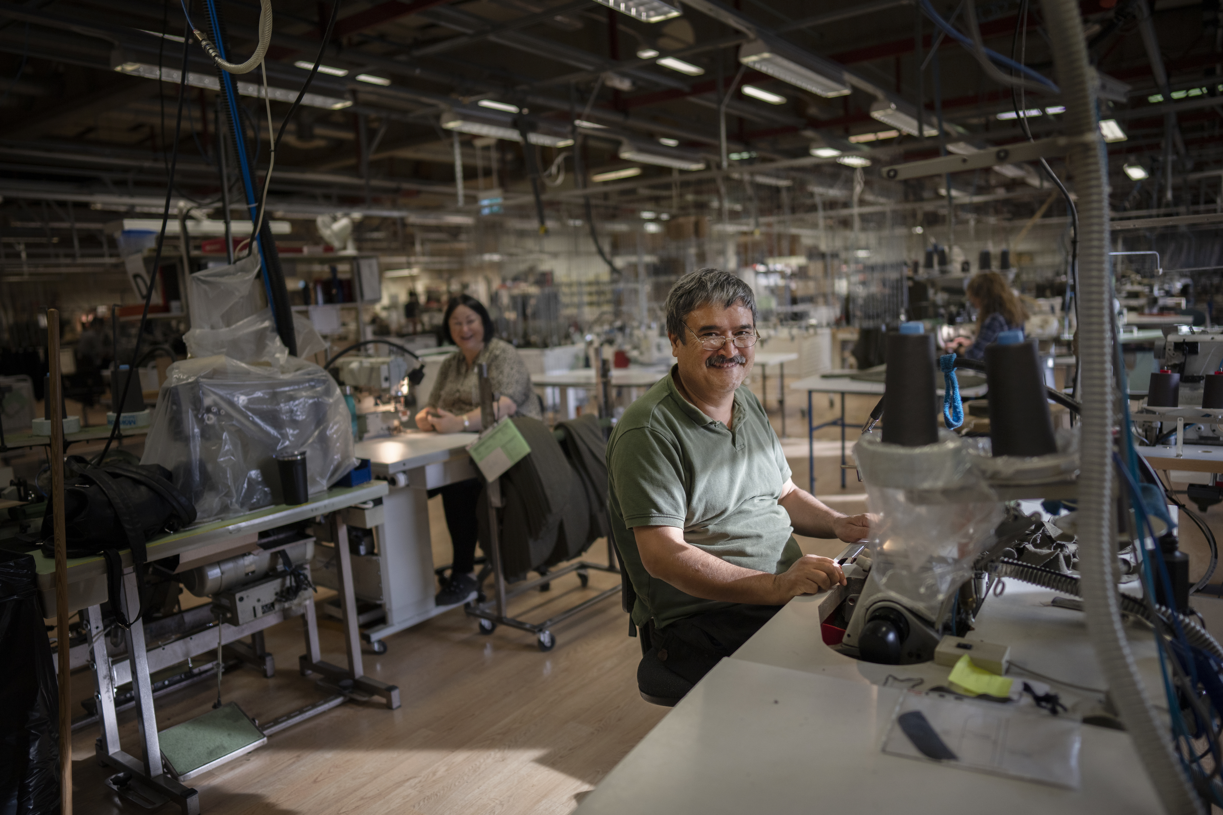 En medelålders man ler vid en symaskin, i bakgrunden skrattar en kollega.