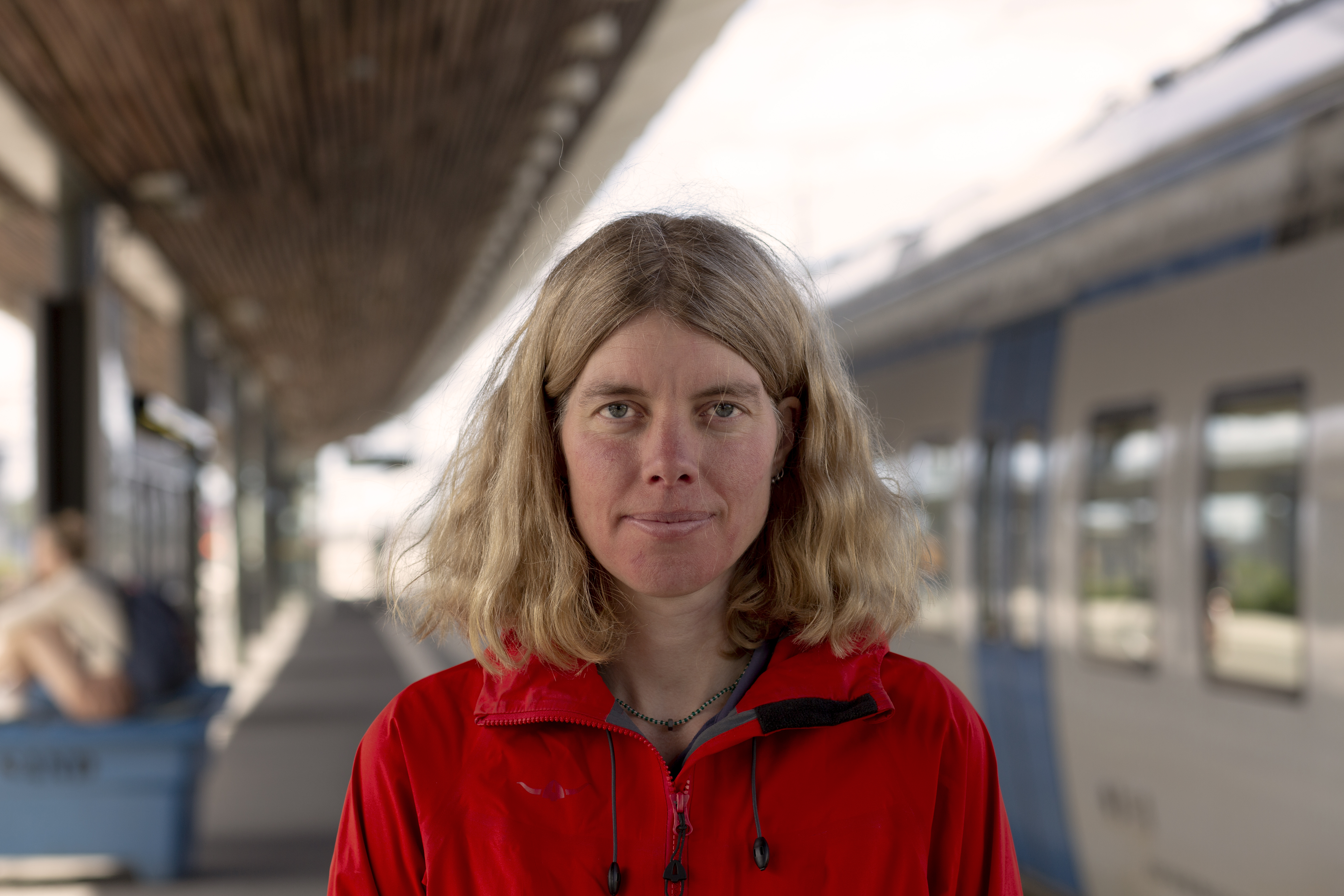 Vänsterpartiets Anna Sehlin i röd jacka på en pendeltågsstation framför ett vitt pendeltåg med blå dörrar.