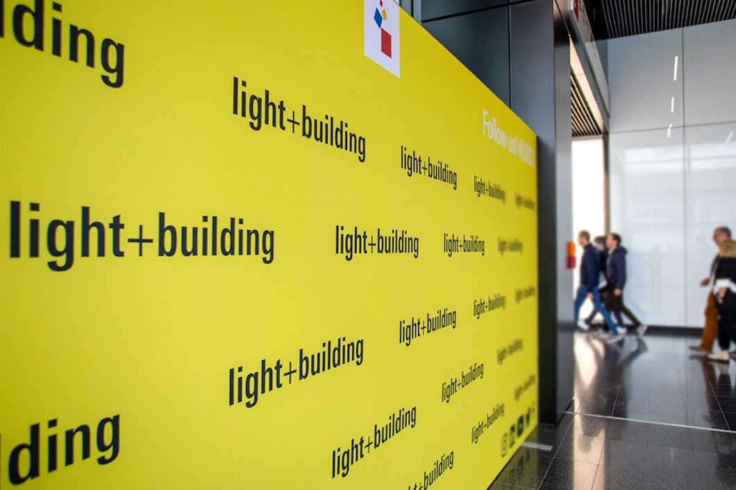 Gul vägg med lesten "light+building" upprepade gång, och bredvid går några människor.