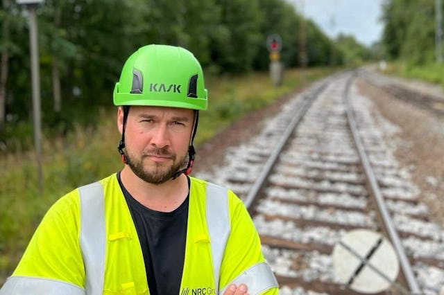 Johan Mähler med armarna i kors och en allvarlig min på ett tågspår. Han har på sig en gul varselväst och en grön hjälm.