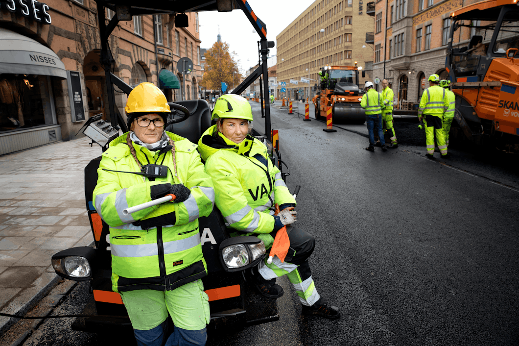 Signalvakterna Annica Björk och Jeanette Lundberg i full mundering av gula varselkläder vid en gata där asfalten läggs om. De står lutade mot en asfaltsmaskin.