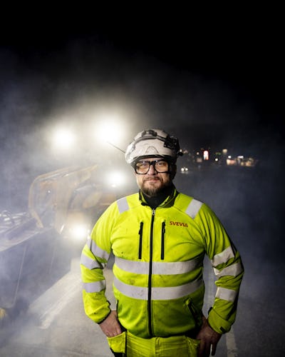 Bill Nylander tittar in i kameran med asfaltmaskinerna i bakgrunden under pågående asfalteringsarbete i mörkret.