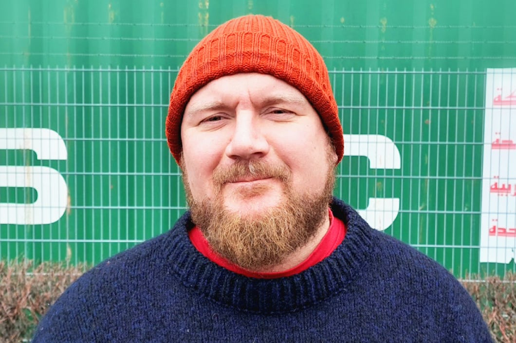 Erik Helgeson med skägg, röd stickad mössa och blå tröja står utomhus framför ett grönt staket och en container av metall.