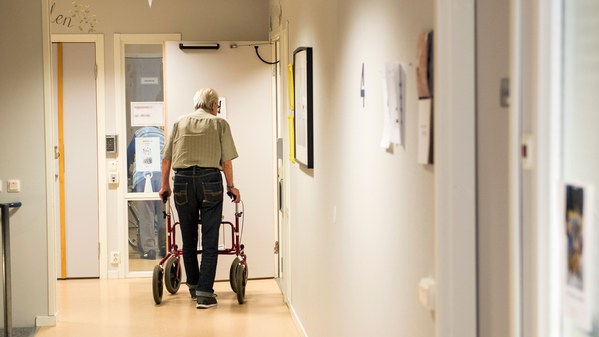Gammal man med rullator i korridor på äldreboende.