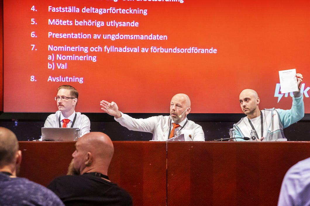 Några män sitter på en scen, framför publik, och en av dem pekar med hela handen.