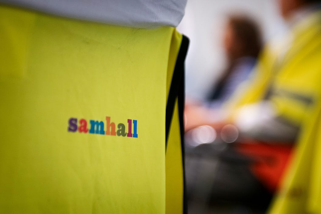 Närbild på en varselväst med texten "Samhall". I den suddiga bakgrunden sitter människor, vilket antyder ett möte eller gruppaktivitet.
