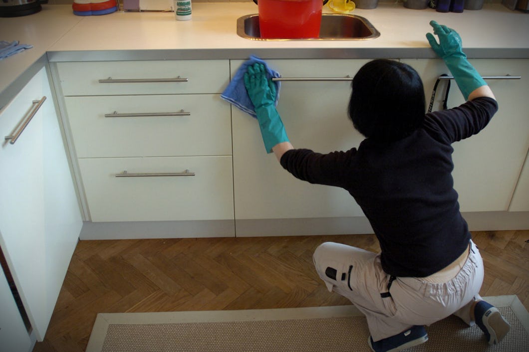 En person med handskar sitter på knä på golvet och torkar av köksluckor med en trasa vid diskbänken.