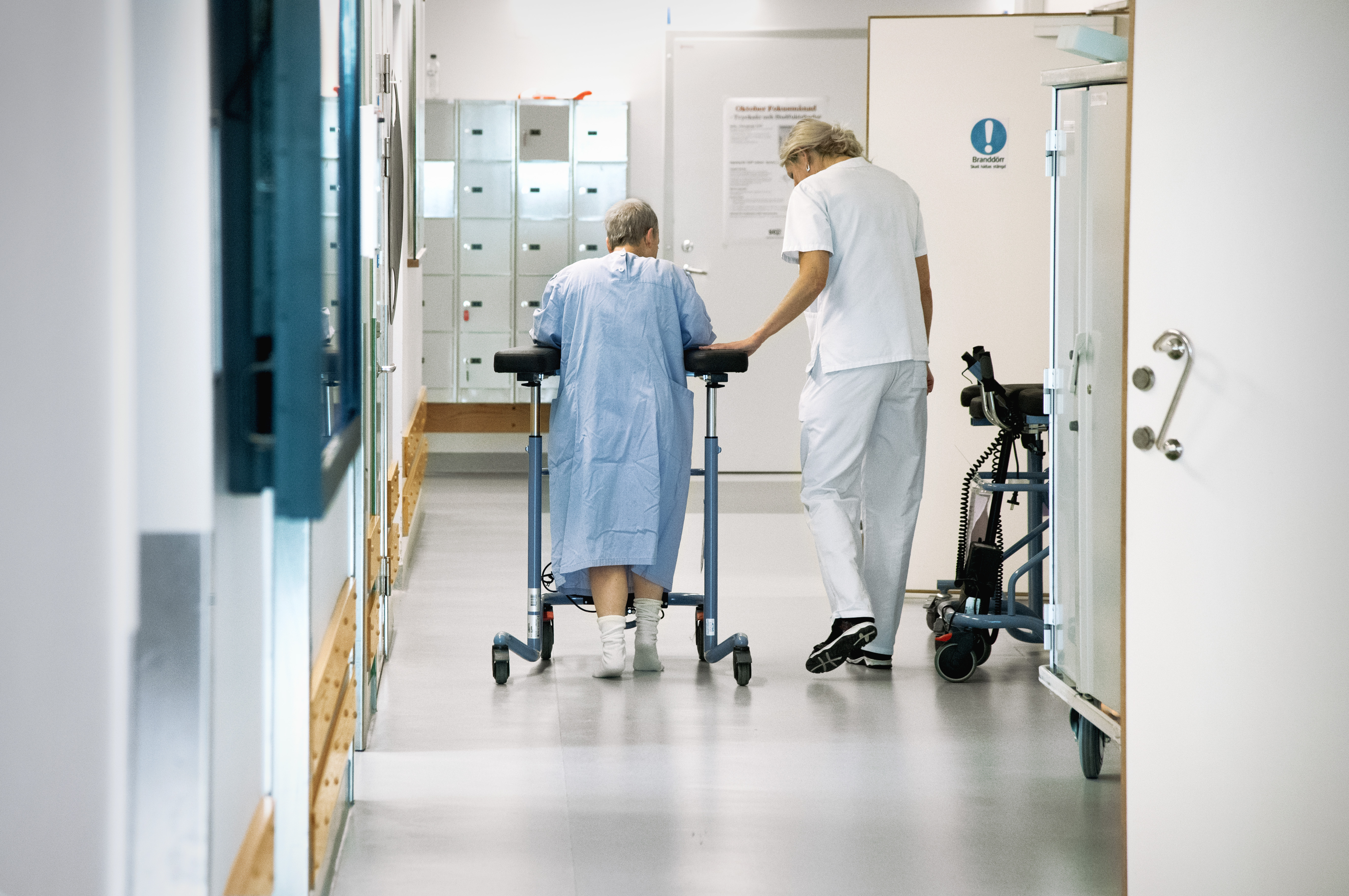 En kvinnlig patient med en rollator får hjälp av personal att ta sig fram i en korridor på ett sjukhus