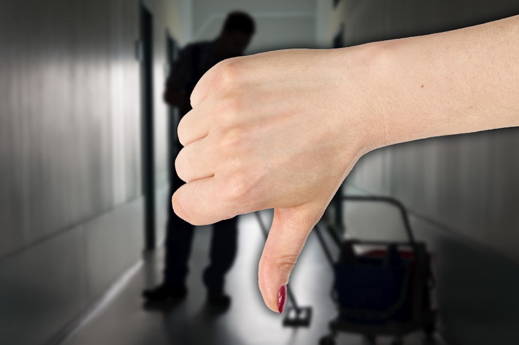 En hand gör en tumme ner-gest i förgrunden. I bakgrunden står en person i en korridor med städutrustning.