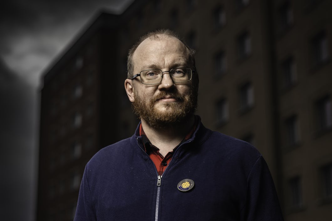 Kim Nilsson har glasögon och skägg och står framför en mörk byggnad.