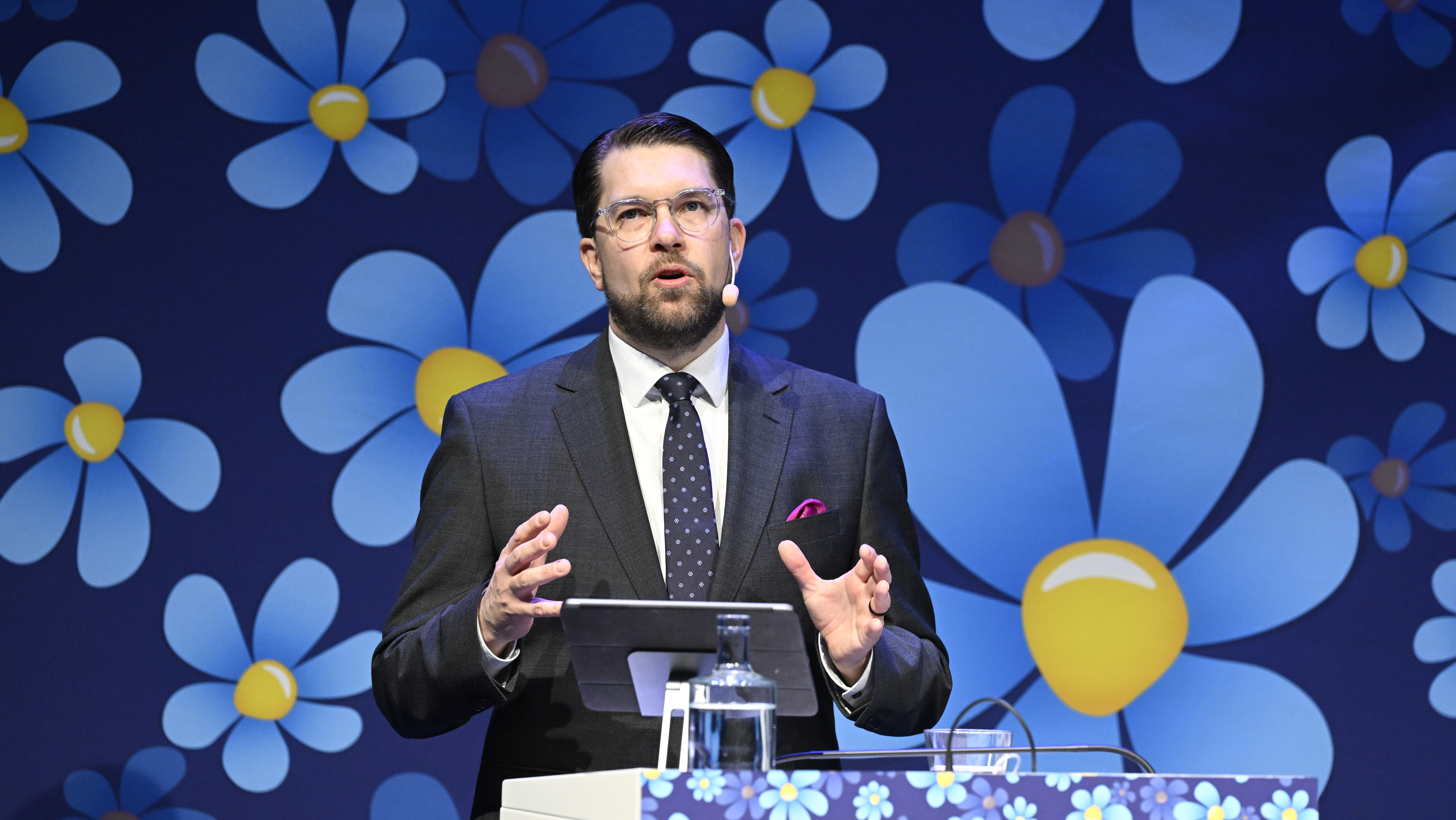 En man i kostym som står framför en blå blommabakgrund.