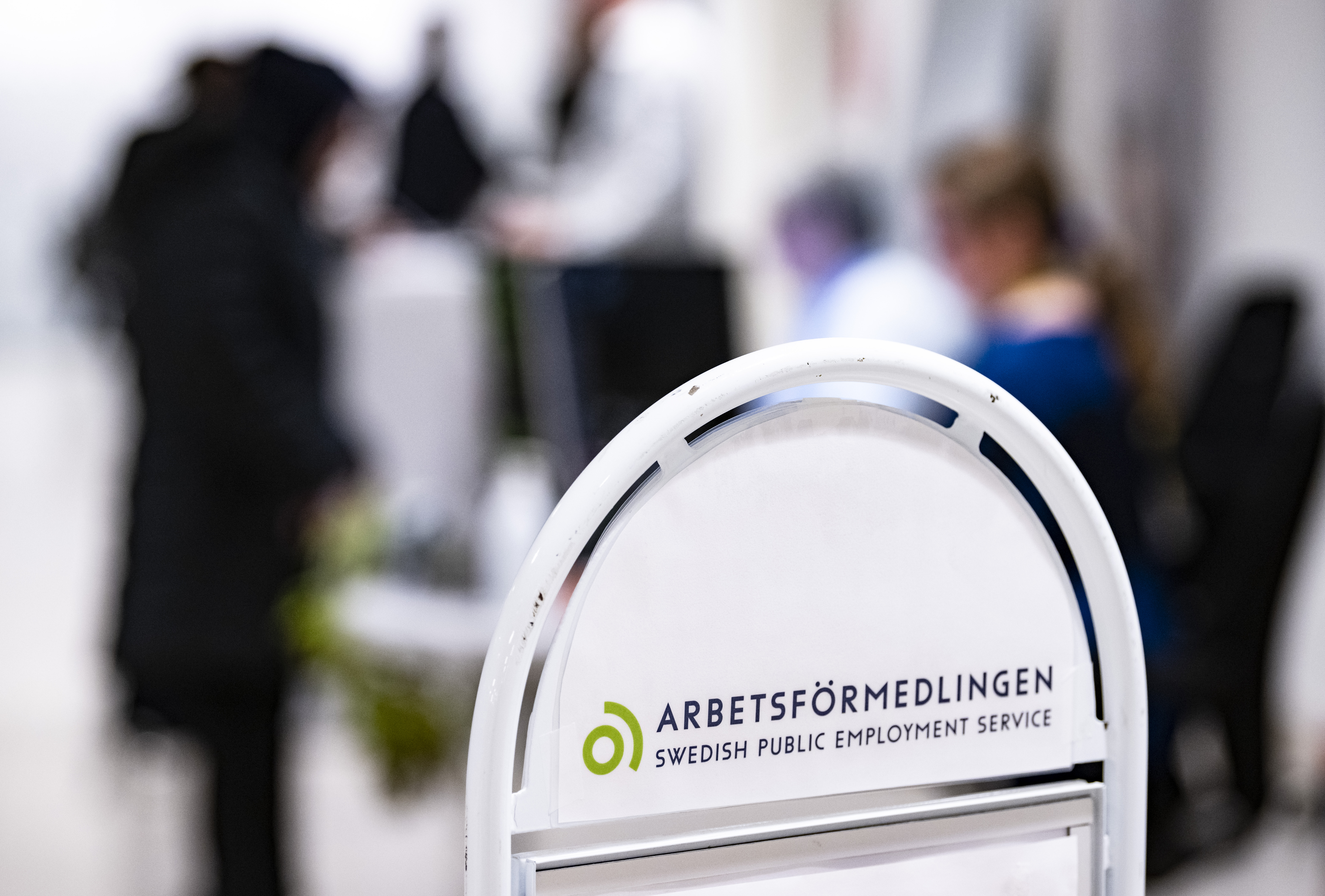 En vit skylt där det står Arbetsförmedlingen framför en grupp människor som syns suddigt i bakgrunden.