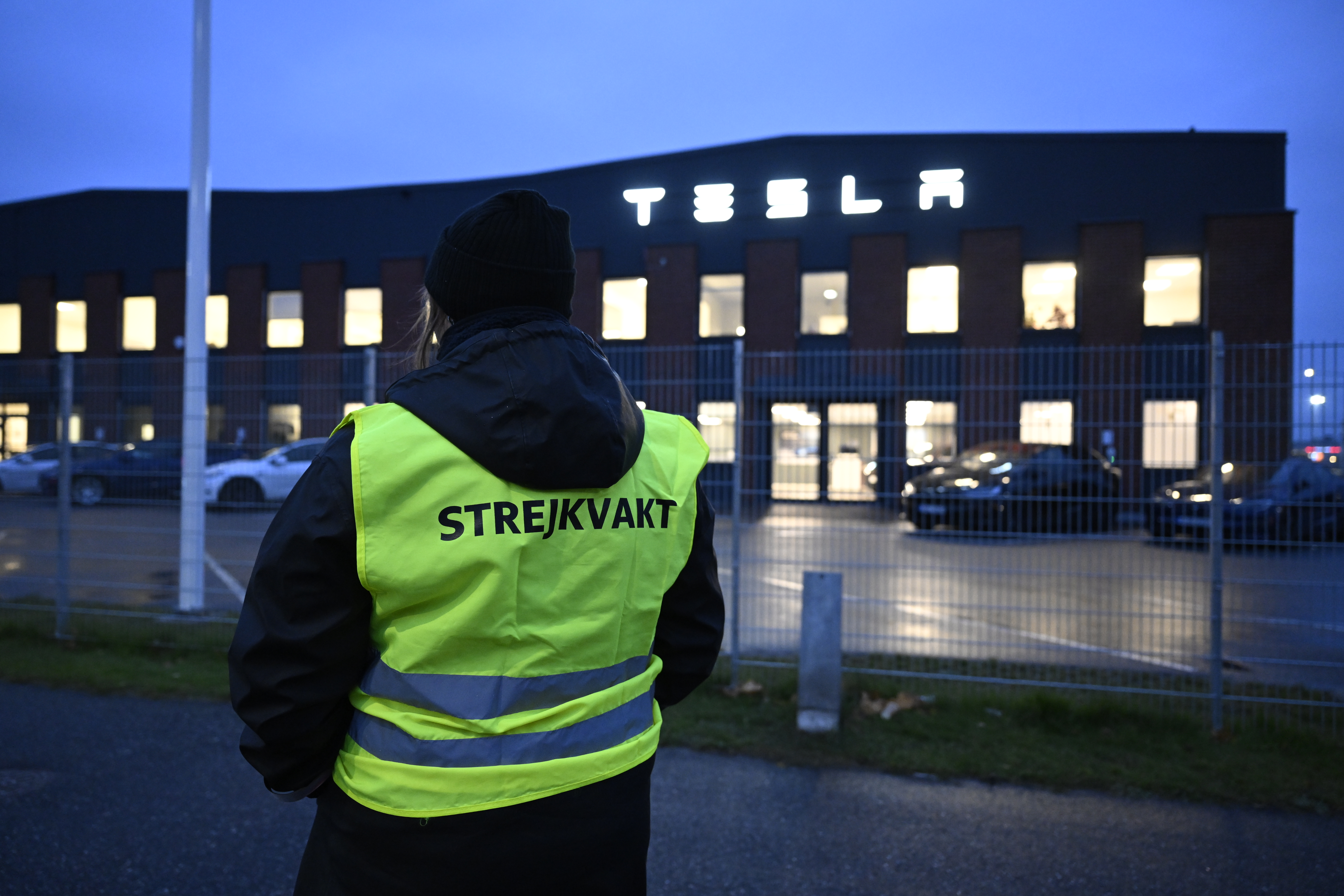 En strejkvakt i en gul väst står med ryggen vänd mot kameran framför en byggnad med texten ”Tesla” på fasaden.