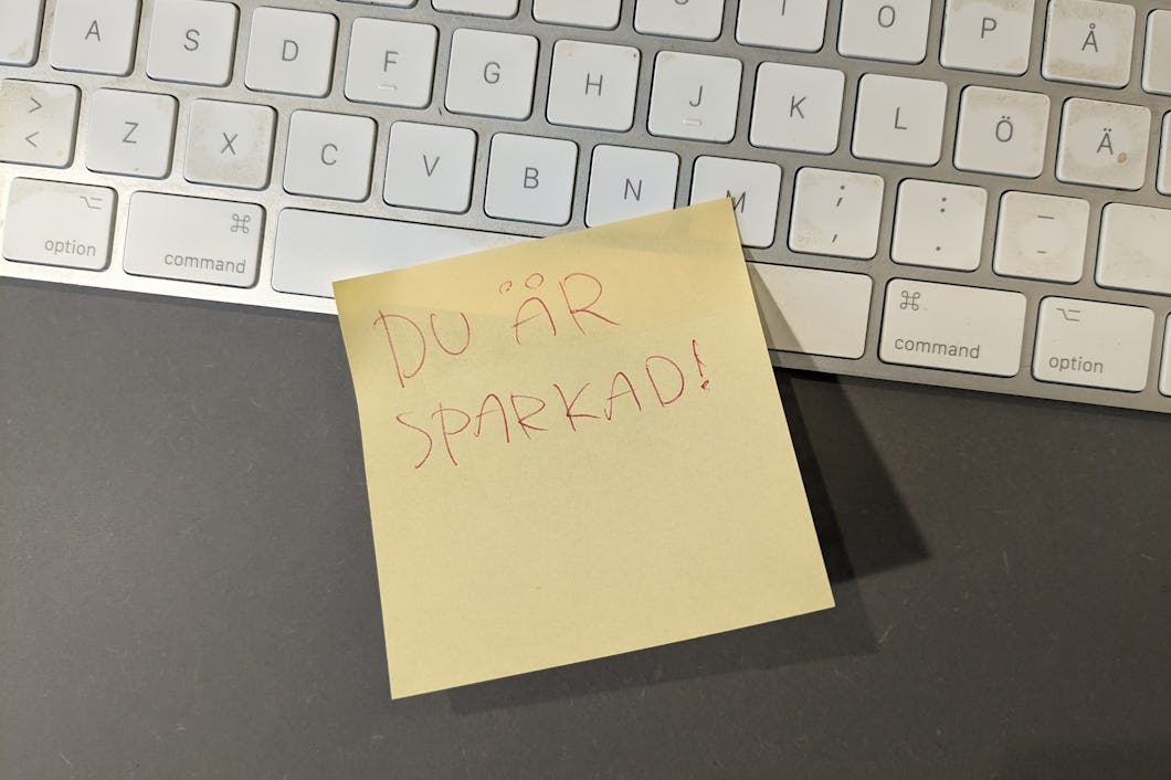 En klisterlapp med frasen "du är sparkad!" handskriven på den, placerad på en grå yta bredvid ett vitt datortangentbord.