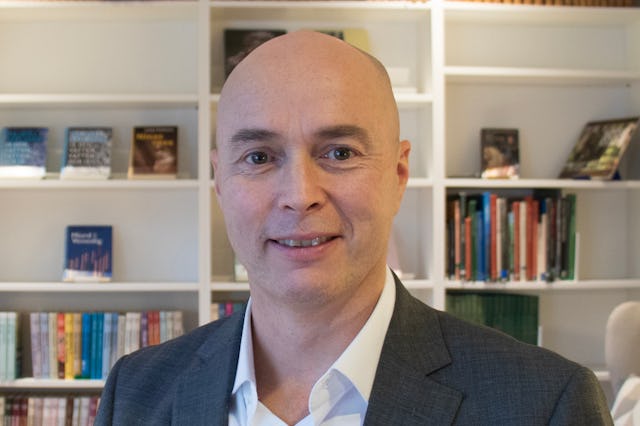 En skallig man i kostym som står framför en bokhylla.