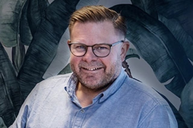 En man i en blå skjorta som står framför en tropisk vägg.